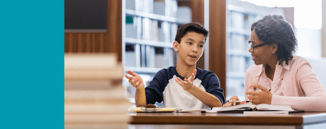 Middle school student speaking with a teacher in a library.